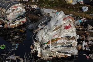 Large piles of compressed waste materials are seen near a polluted water source in an urban setting, highlighting environmental concerns photo