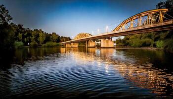 Bridge over a wide river at dusk, lights beginning to illuminate its arches. Reflections shimmer on the rippling water. Trees line the riverbanks. A tranquil and scenic view at twilight. photo