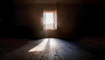 Dark, empty room with sunlight streaming through an open window, casting a bright beam across the wooden floor. A view of the outside landscape. Evokes feelings of hope and solitude. photo