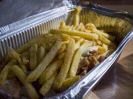 French fries piled on a kebab drizzled with sauce, all nestled in an aluminum takeaway box resting on a wooden table, creating a tempting fast food meal photo