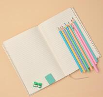 Top view of an open notebook with pastel-colored pencils, green sharpener, and turquoise eraser on a beige background. photo