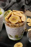Two glasses of chia seed pudding, one topped with diced kiwi and the other with granola and banana chips, are on a dark surface with ingredients. Above photo