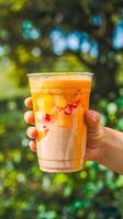 A close-up image of a vibrant mango smoothie in a hand, with a natural blurred green background, conveying freshness, tropical vibes, and summer relaxation. photo
