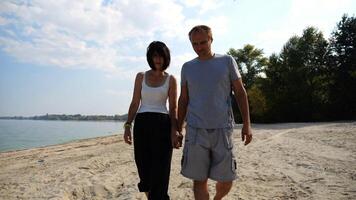 Romantic couple holding hands of each other and walking at coastline. Happy pair going along seashore at sunny day. Lovers spending time together at resort. Concept of vacation or holiday. Slow motion photo