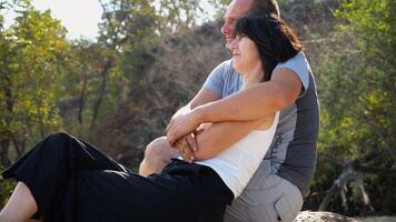 Happy pair hugs during sitting on wooden log on beach. Romantic couple admiring sea landscape at sunny day. Lovers spending time together at resort. Concept of summer vacation or holiday. Slow mo photo