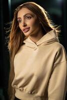 young woman long flowing hair poses in soft beige hoodie smiling gently while standing in intimate indoor setting dim lighting creating atmospheric effect photo