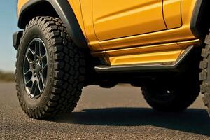 Close-up side view of a yellow off-road vehicle with aggressive tread tires and black alloy wheels, emphasizing rugged build and outdoor readiness on a sunlit road. photo
