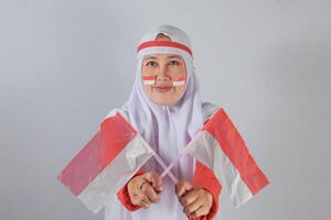 Determined Southeast Asian Muslim woman in red and white hijab holding two small red and white flags crossed forward with face paint showing national pride and unity photo