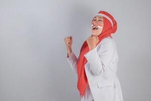 Joyful Indonesian woman celebrating with clenched fists and patriotic face paint expressing national pride and energetic triumph emotion in victory pose perfect for themes of motivation and unity photo