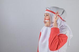 Side view of Indonesian woman in red and white hijab with flag-painted cheek making expressive hand gesture and puckered lips showing playful and proud mood for Independence Day photo