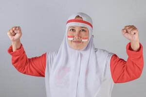 Confident Indonesian Muslim woman in white hijab and red clothing with patriotic face paint raising both fists in strong determined pose symbolizing national pride and empowerment photo