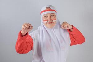 Smiling Indonesian Muslim woman in white hijab and red clothing with patriotic face paint raising both fists in a joyful and confident expression showing national pride and celebration photo