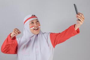 Muslim woman dressed in red and white taking selfie with raised fist and joyful expression, celebrating Indonesian Independence Day with pride and enthusiasm in a digital way photo