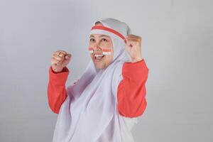 Smiling Indonesian woman in red and white hijab costume raising fists in enthusiastic pose with painted national flag on cheeks to express joy and pride for Independence Day celebration photo