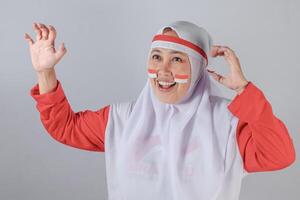 Happy Indonesian Muslim woman in white hijab and red outfit with patriotic flag face paint raising hand and smiling joyfully while calling out or cheering during celebration event photo