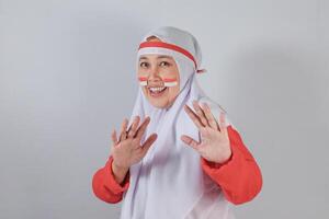 Playful Muslim woman in hijab with Indonesian flag face paint showing both hands in surprised cheerful expression, representing cultural pride and joyful interaction photo