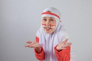 Confused Muslim woman in hijab with Indonesian flag face paint making uncertain gesture with both hands raised, expressing questioning emotion and cultural identity photo