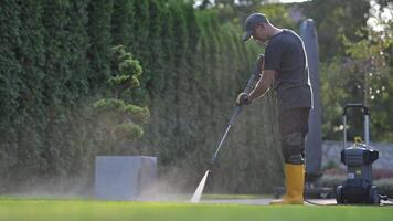 A man in yellow rubber boots and a black shirt is using a pressure washer to clean the video
