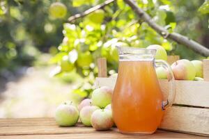 Apples, box of apples and pitcher of fresh apple juice on wooden table with garden background. Harvest of apples. Juice production photo