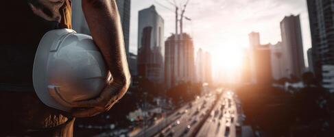 The construction worker holds a helmet while observing the city skyline at sunset. photo