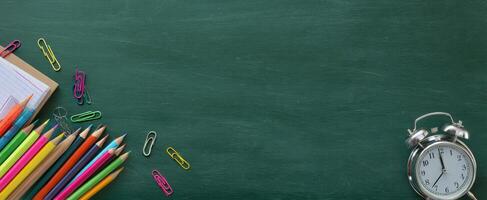 The colorful pencils and alarm clock on a green chalkboard background. photo