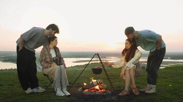 Young people are having pleasant conversation and enjoying themselves while cooking over an open fire, with beautiful sunset as a backdrop. video