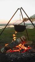 Thick meat soup, cooked over campfire during summer walk, is stirred with wooden spoon. stew is cooked in pot, hanging on hiking tripods, against backdrop of sunset sky and landscape with fields and river. video