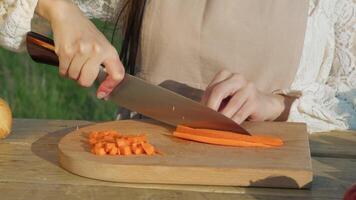 A young woman, dressed in a modest, traditional outfit and wearing a brown apron, is carefully slicing carrots using a sharp knife on a wooden cutting board, preparing for dinner. video