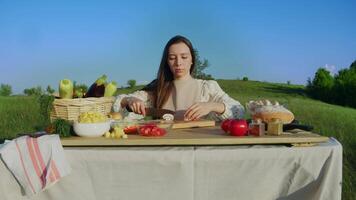 Seated at table in midst of nature, young woman in dress and apron meticulously slices mushrooms into bite-sized pieces, surrounded by plethora of vegetables, preparing for summer feast. video