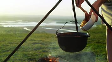 While cooking over campfire, cook hangs pot on tripod and adds diced potatoes to soup or stew, against backdrop of summer landscape. video