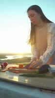 Cook carefully cuts carrots into thin slices. vegetables are laid out on table, surrounded by other fresh and mouth-watering products, against backdrop of breathtaking sunset, in heart of nature. video