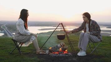 Women are chatting while cooking soup over open fire, surrounded by beautiful scenery of summer countryside. They are sitting on camping chairs and enjoying each other's company. video