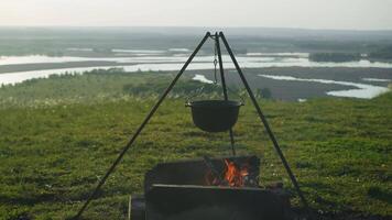 Cast-iron tripod with metal pot rests over campfire on hillside on summer's day. Vegetable stew or soup is being prepared in it over open fire as light meal for hikers. video