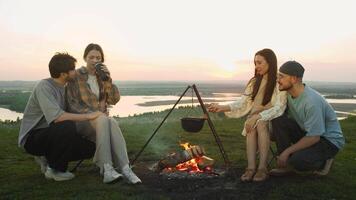 Young couples are relaxing outdoors, sitting around a campfire as they cook food. One girl is using a ladle to scoop some soup from a pot, trying to see if it's ready to eat. video