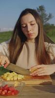 A young woman dressed in peasant clothes is sitting in a rural area, in a field, at a table and cutting potatoes for dinner on a wooden cutting board. Vertical. Portrait. video