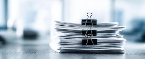 The stack of documents secured with binder clips resting on a wooden desk. photo