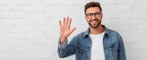 The smiling man in denim jacket waves cheerfully against a white wall. photo