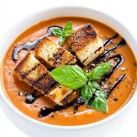 A close-up of a bowl of creamy tomato basil soup, topped with grilled cheese croutons and a drizzle photo