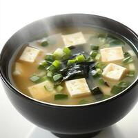 A close-up shot of a steaming bowl of miso soup, garnished with tofu cubes, seaweed, and green onions, offering a comforting Japanese appetizer. The miso soup, with its savory umami flavor and light photo
