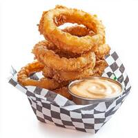 A stack of crispy, golden-brown onion rings, served with a side of creamy dipping sauce, resting on a checkered paper-lined basket. The onion rings, with their sweet, caramelized flavor and satisfying photo
