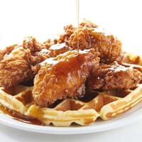 A detailed close-up of a plate of chicken and waffles, featuring crispy fried chicken and fluffy waffles, drizzled with maple syrup. The chicken and waffles, with their contrasting textures and photo