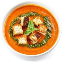 An image of a bowl of creamy tomato soup, topped with grilled cheese croutons and a swirl of pesto, presented on a white background. The creamy tomato soup, with its rich tomato flavor and smooth photo