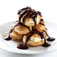 A close-up of a profiterole au chocolat, a cream puff filled with vanilla pastry cream and topped with chocolate sauce, arranged in a stack on a plate, showcasing the light and airy pastry and the photo