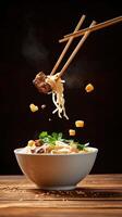 Noodles with meat held by chopsticks, served in bowl on wooden table. Background blurred. Stock photo