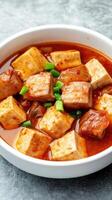 Tofu cubes with a rich, savory sauce in a white bowl on gray surface. Food photography photo