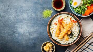 Udon noodles with tempura shrimp and veggies, served on a textured, grey background. Possible food blog use photo