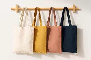 Four canvas tote bags in neutral and warm tones hanging on wooden wall hook against white background in natural light photo