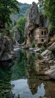 Ruined stone building beside tranquil river surrounded by rocky cliffs and lush greenery under an overcast sky photo