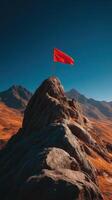 Red flag stands tall on rocky peak against clear blue sky and mountain backdrop during bright daylight hours photo