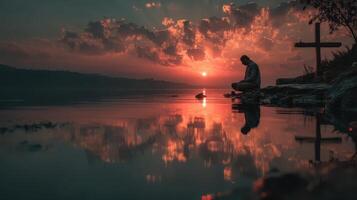 Man meditating by the water at sunset with a cross in the background photo
